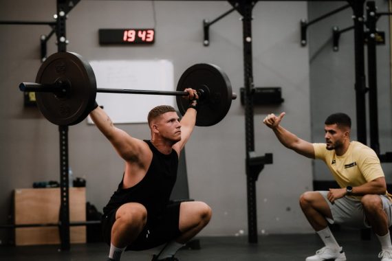 A muscular man performing a weightlifting squat with a coach guiding him indoors.